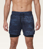 A man wearing navy blue camouflage swim trunks with a black drawstring and a black inner liner, shown from the waist to mid-thigh against a light beige studio background.