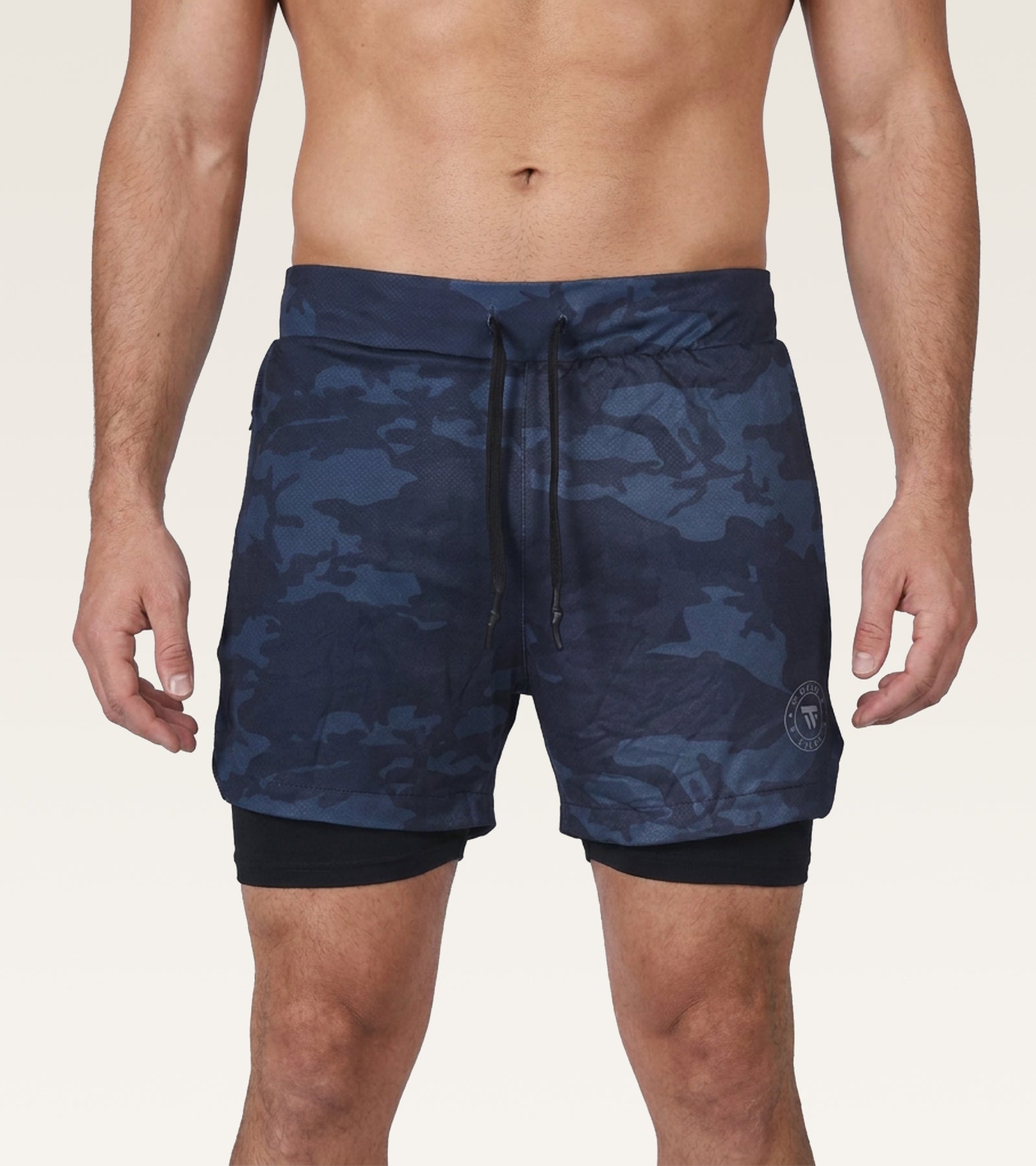 A man wearing navy blue camouflage swim trunks with a black drawstring and a black inner liner, shown from the waist to mid-thigh against a light beige studio background.