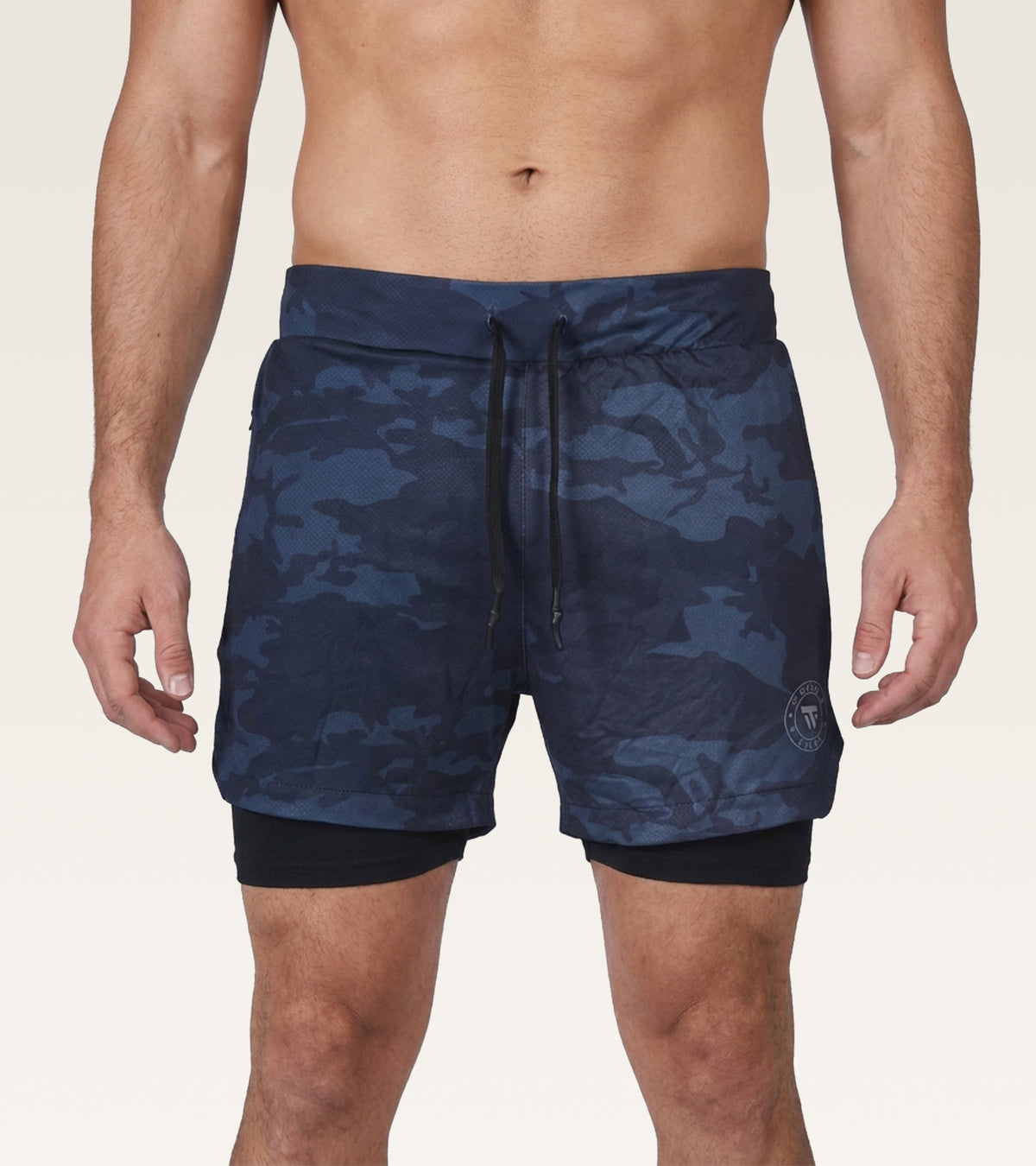A man wearing navy blue camouflage swim trunks with a black drawstring and a black inner liner, shown from the waist to mid-thigh against a light beige studio background.