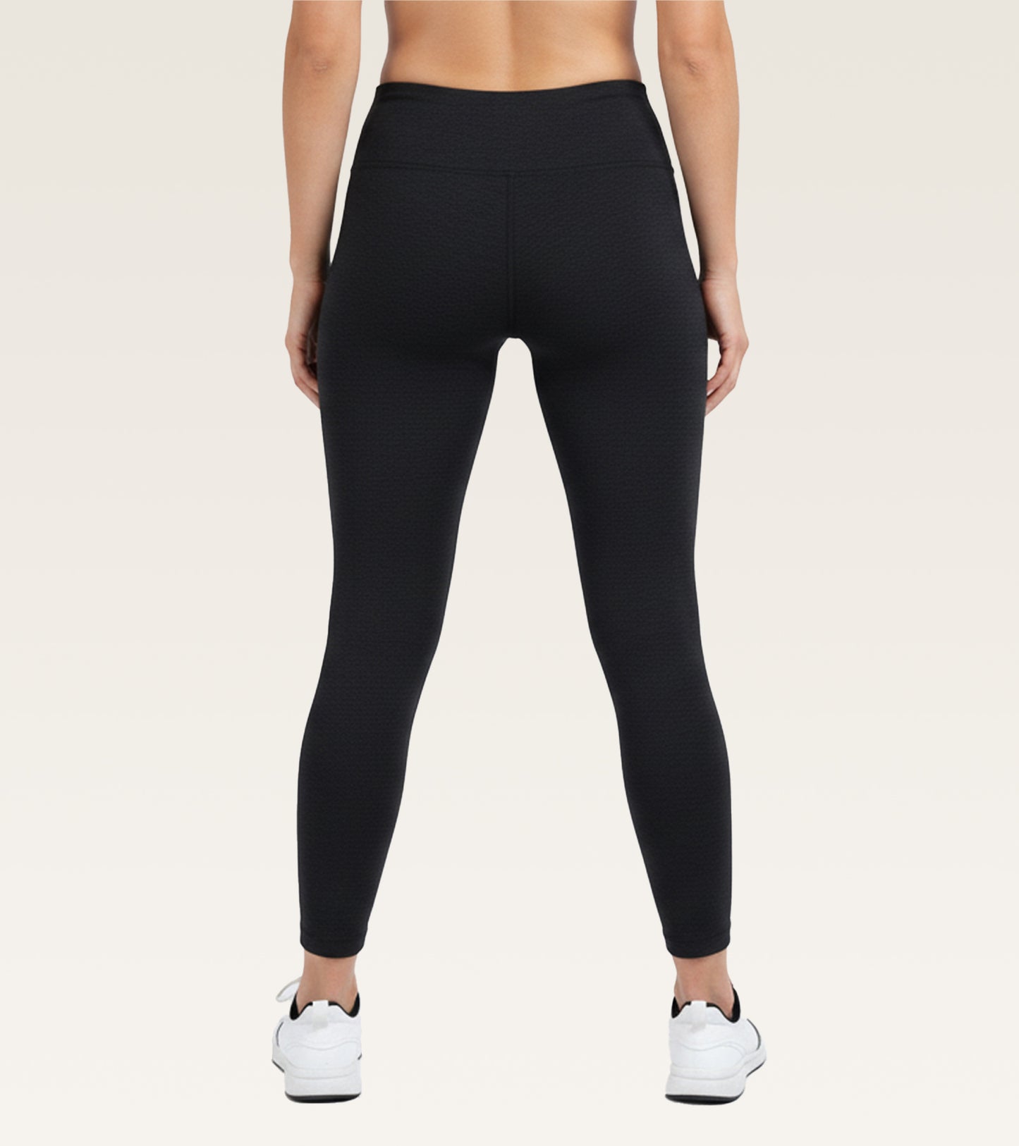 Rear view of a model wearing black high-waisted leggings and white sneakers against a light beige studio background.