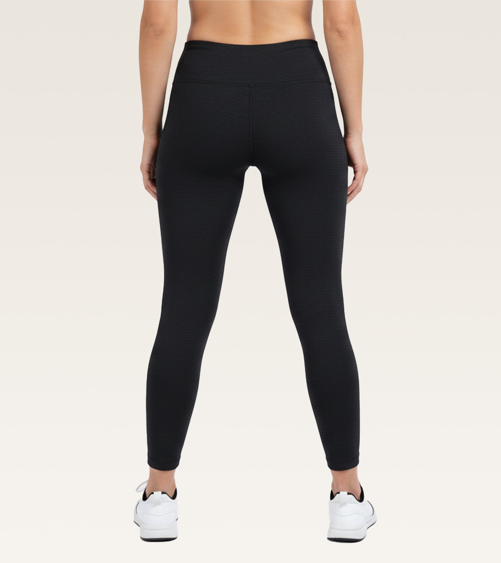 Rear view of a model wearing black high-waisted leggings and white sneakers against a light beige studio background.