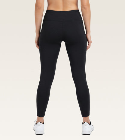 Rear view of a model wearing black high-waisted leggings and white sneakers against a light beige studio background.